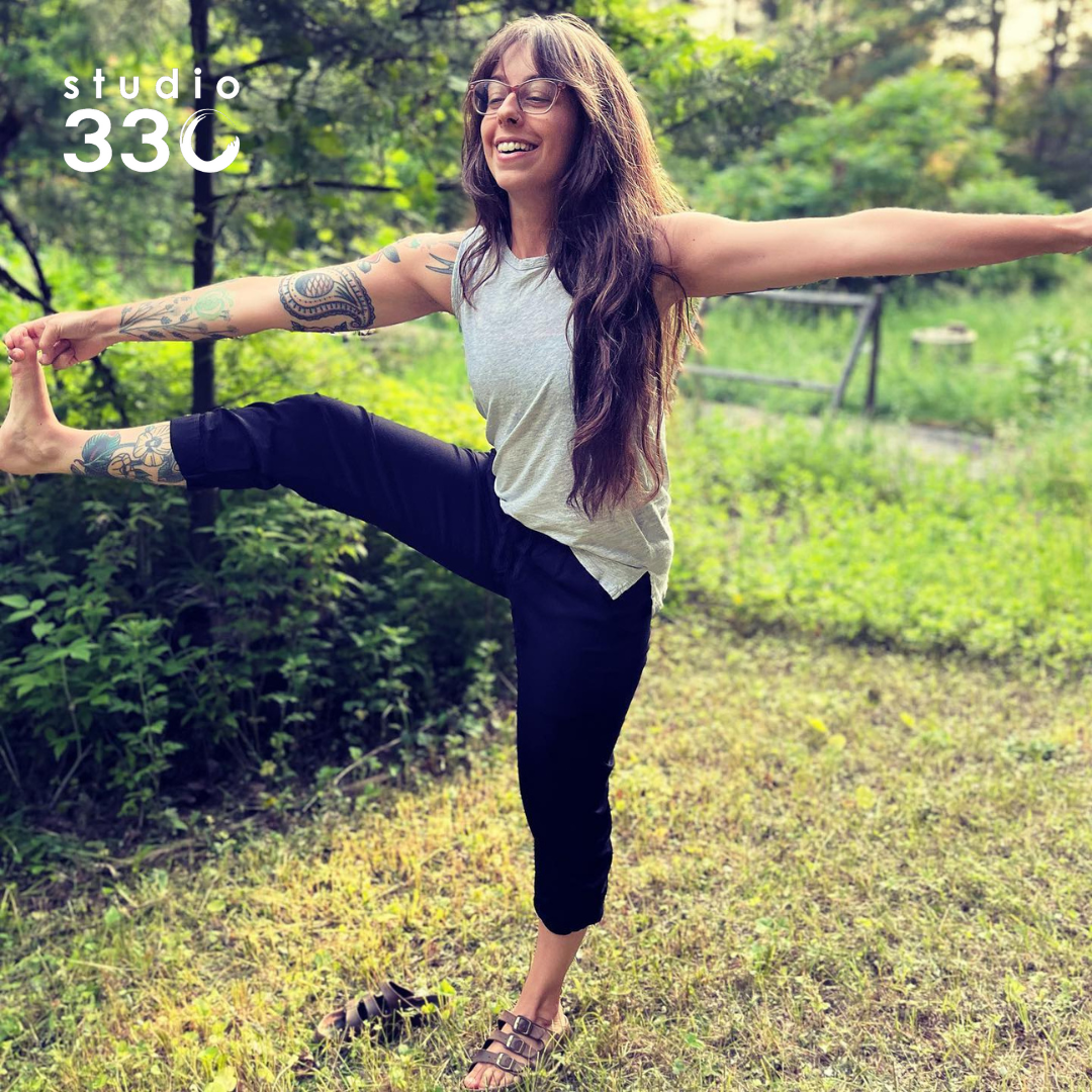 Chelsea standing on one foot outdoors, performing a toe hold with a smile