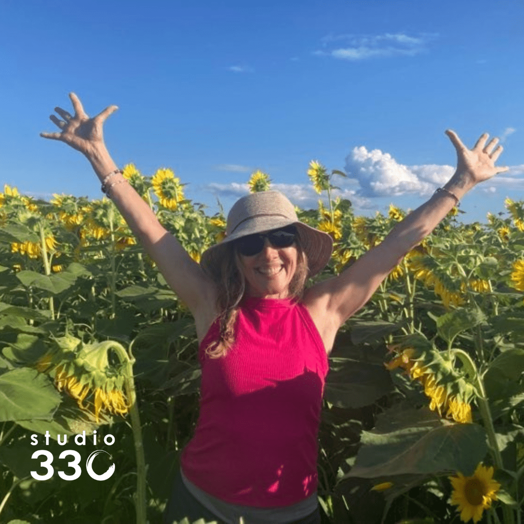 Lori with her hands up, smiling, standing in a field of sunflowers